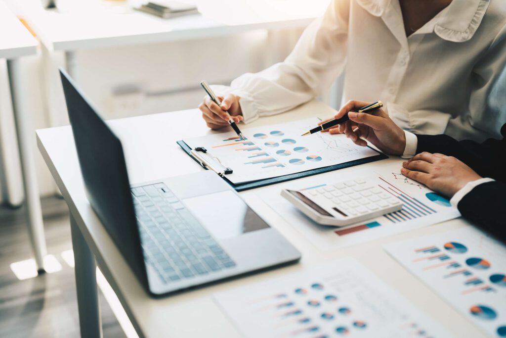 Zwei Personen analysieren Berichte auf einem Tisch mit Laptop und Diagrammen.